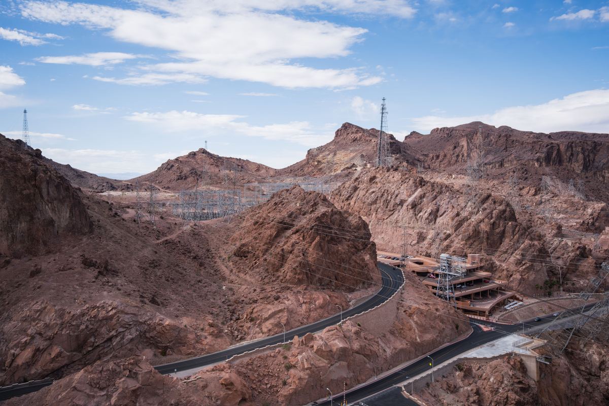 A paved road curves through rugged red-brown rocky hills, with a power switchyard, pylons and a tiered structure under a blue sky with clouds.